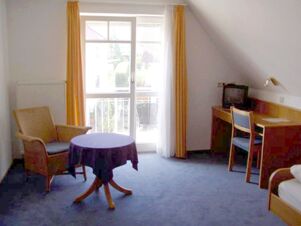 Helles G&auml;stezimmer im Gasthof Schwarzer Adler mit blauem Teppich, kleinem Tisch, Stuhl und Schreibtisch vor einem Balkonfenster mit gelben Vorh&auml;ngen.