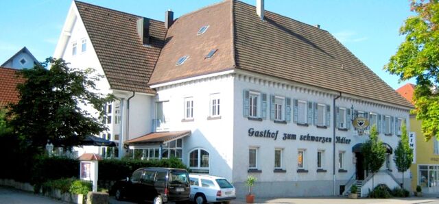 Au&szlig;enansicht des Gasthofs Schwarzer Adler mit traditionellem wei&szlig;em Geb&auml;ude, blauen Fensterl&auml;den und Parkpl&auml;tzen davor an einem sonnigen Tag.