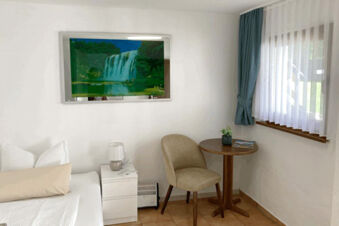 Small seating area with beige chair, round wooden table, potted plant, curtains, and waterfall picture in Pension Lachmann room