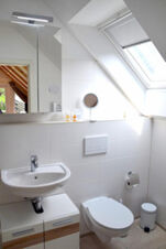 Bright white bathroom in Pension Lachmann with a sink, wall-mounted toilet, mirror cabinet, skylight window, and small trash bin