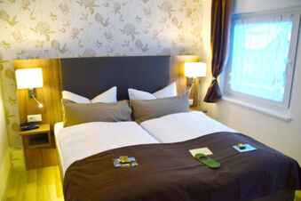 Double bed with white and brown bedding, two brown pillows, nightstands with lamps, floral wallpaper, and a window at Pension Lachmann