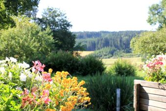 Bunte Blumen und gr&uuml;ne Pflanzen in einem Garten mit Wald und Feldern im Hintergrund beim Landgasthof Haueis an einem sonnigen Tag.