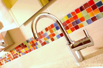 Moderner Wasserhahn vor bunter Mosaikfliesenwand in einem Badezimmer im Landgasthof Haueis mit hellem Waschbeckenbereich.