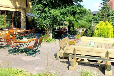 Au&szlig;enbereich mit Holztischen und St&uuml;hlen im Garten vom Landgasthof Haueis, umgeben von B&auml;umen und Bepflanzungen bei Sonnenschein.