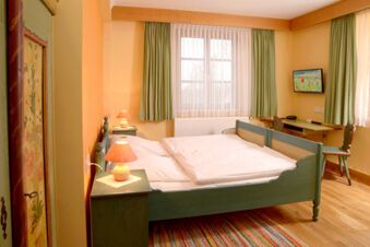 Schlicht eingerichtetes Zimmer mit Doppelbett, Holzm&ouml;beln und gr&uuml;nen Vorh&auml;ngen im Landgasthof Haueis mit warmem Lichtakzent.