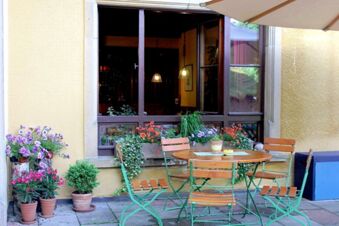 Gem&uuml;tlicher Au&szlig;enbereich mit Holztisch, St&uuml;hlen und vielen bl&uuml;henden Pflanzen vor dem Landgasthof Haueis an einer gelben Hauswand.