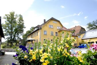 Au&szlig;enansicht von Landgasthof Haueis mit gelbem Geb&auml;ude, bl&uuml;henden Blumen im Vordergrund und Parkplatz im Hintergrund bei sonnigem Wetter.