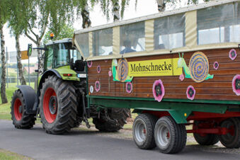 Ein Traktor mit dem farbenfrohen Anh&auml;nger 'Mohnschnecke' steht auf einem Weg vor B&auml;umen, angeboten vom Landhotel-Restaurant Mei&szlig;nerhof.