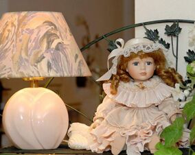 Porcelain doll dressed in vintage pink lace outfit sitting on a metal shelf next to a lit floral-pattern lamp at Landhaus Heide Romantisches Heidehotel Garni Nichtraucher