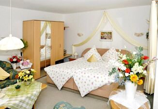 Bedroom with double bed covered in floral bedding, wooden wardrobe with mirrors, sofa, and flower arrangements at Landhaus Heide Romantisches Heidehotel Garni Nichtraucher