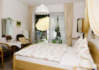 Bedroom with floral bedding and pillows, wooden furniture, a small table with chairs, and open doors to a patio with a yellow umbrella at Landhaus Heide Romantisches Heidehotel Garni Nichtraucher