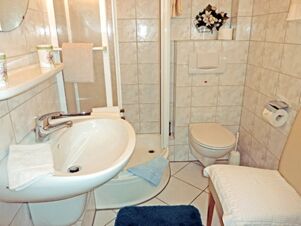 Bathroom with white sink, shower cabin, toilet, beige chair and blue rug at Landhaus Heide Romantisches Heidehotel Garni Nichtraucher