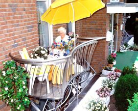 Elderly woman sitting at a table with flowers under a yellow umbrella on a balcony at Landhaus Heide Romantisches Heidehotel Garni Nichtraucher