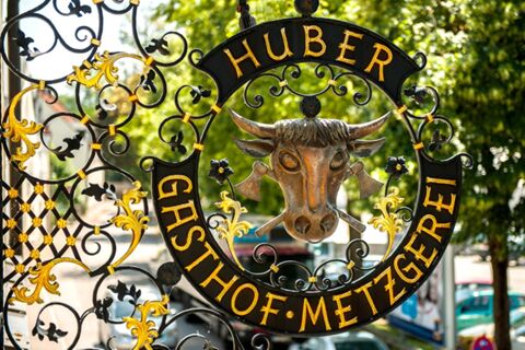 Detailaufnahme eines kunstvoll verzierten Eisenschilds mit goldenem Schriftzug und Kuhkopf, markiert mit Hotel Gasthof Huber im Freien.