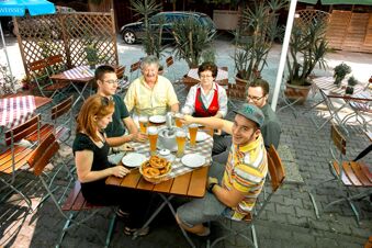 Sechs Menschen sitzen gemeinsam an einem Tisch mit Bier und Brezeln im Au&szlig;enbereich des Hotel Gasthof Huber bei Sonnenschein.