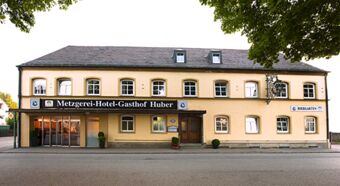 Die Au&szlig;enansicht des Hotel Gasthof Huber zeigt ein zweist&ouml;ckiges Geb&auml;ude mit traditionellen Fenstern und einer Beschilderung inklusive Biergarten und Metzgerei.