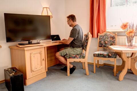 Un bărbat lucrează la un laptop la o masă de lemn &icirc;ntr-o cameră de hotel confortabilă, cu televizor și decorațiuni florale, la Hotelul Gasthof Huber.