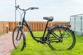 Ein schwarzes Fahrrad steht auf einer Rasenfl&auml;che neben einem gepflasterten Weg, umgeben von einem Holzzaun bei Meyenburg & Gerds H&ouml;ft Appartements am Wattenmeer.