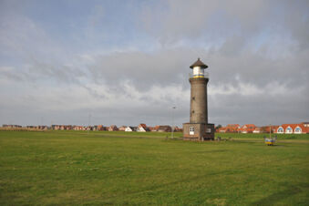 Weitl&auml;ufige gr&uuml;ne Wiese mit einem historischen Leuchtturm und H&auml;usern im Hintergrund bei Meyenburg & Gerds H&ouml;ft Appartements am Wattenmeer.