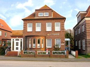 A brick two-story building with a tiled roof, white framed windows, a small garden with a fence, and a sign for Marinas Ferienwohnungen