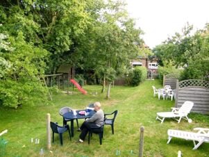 A garden area at Marinas Ferienwohnungen with a woman sitting at a black table, playground slide, and white lounge chairs on grass