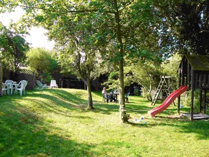 A sunny garden at Marinas Ferienwohnungen with green trees, a red slide, plastic chairs, tables, and a sandbox with scattered toys
