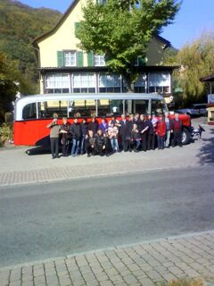 Vor dem Hotel-Restaurant zum Wilden Mann steht eine gro&szlig;e Gruppe von Menschen neben einem historischen roten und wei&szlig;en Bus vor einem gelben Geb&auml;ude mit gr&uuml;nen Fensterl&auml;den.