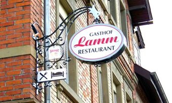 Gasthof Lamm Hotel und Restaurant sign showing a white oval with red and black letters attached to a brick building with additional symbols for dining and lodging