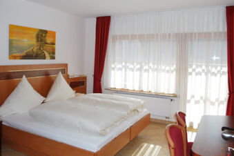 Double bed with white bedding, red curtains, sheer white window drapes, two chairs, and a desk in Gasthof Lamm Hotel und Restaurant room