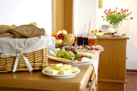 Comptoir du petit-d&eacute;jeuner avec du pain frais, de la charcuterie, des fruits, de la confiture et du beurre dans la salle &agrave; manger de la pension Garni Zweck avec un arrangement floral.