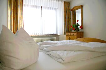 Double bed with white bedding near a window and wooden dresser with mirror and flowers in Pension Garni Zweck
