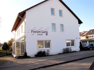 White two-story building with several windows and a sign for Pension Garni Zweck on the side near parked cars