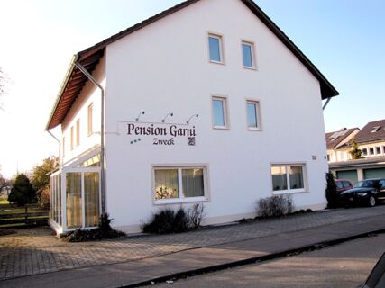 White two-story building with several windows and a sign for Pension Garni Zweck on the side near parked cars