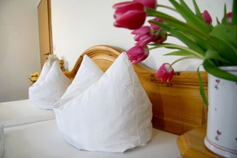 Chambre &agrave; la d&eacute;coration claire avec des coussins blancs, un cadre de lit en bois et un bouquet de fleurs dans le vase de la Pension Garni Zweck.