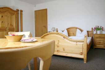 Wooden double bed with white pillows and blankets, wooden wardrobe, table with bowl of apples, and chair at Pension Garni Zweck