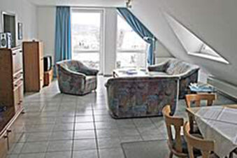 Ger&auml;umiges Wohnzimmer mit Sofa, Sesseln und Essecke in einer Ferienwohnung des G&auml;stehaus Therme-Blick mit gro&szlig;em Fenster und Fliesboden.