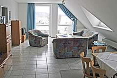 Ger&auml;umiges Wohnzimmer mit Sofa, Sesseln und Essecke in einer Ferienwohnung des G&auml;stehaus Therme-Blick mit gro&szlig;em Fenster und Fliesboden.