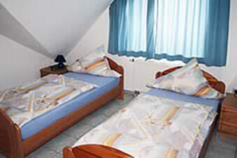 Zweibettzimmer mit Holzm&ouml;beln, Tageslicht durch blaue Gardinen und gemusterte Bettw&auml;sche im Ferienwohnungen G&auml;stehaus Therme-Blick.
