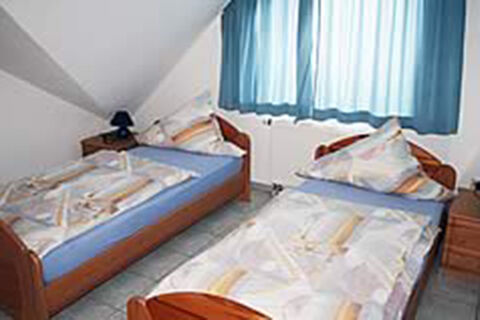 Zweibettzimmer mit Holzm&ouml;beln, Tageslicht durch blaue Gardinen und gemusterte Bettw&auml;sche im Ferienwohnungen G&auml;stehaus Therme-Blick.