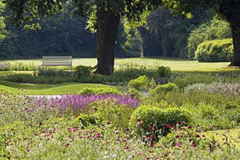 Blumenbeet mit bunten Blumen, gr&uuml;ner Wiese und Baum, davor eine wei&szlig;e Bank im Garten von Ferienwohnungen G&auml;stehaus Therme-Blick.