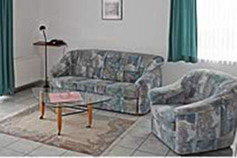 Wohnzimmer mit gemustertem Sofa, Sessel, Glas-Couchtisch und Fenstervorh&auml;ngen in Ferienwohnungen G&auml;stehaus Therme-Blick.