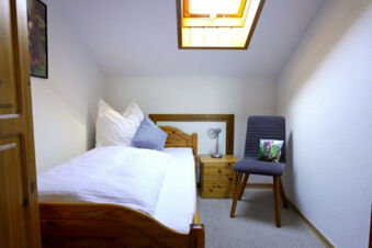 Single wooden bed with white linens and pillows beside a nightstand and a gray chair in Bauernhof Ferienhof K&uuml;hberg bedroom