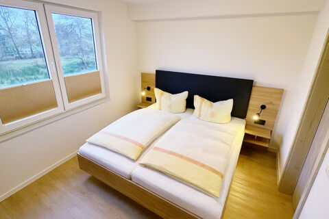 Double bed with beige and white bedding, wooden floors, two bedside tables with reading lamps, and windows at Fewos Haus am W&auml;ldchen