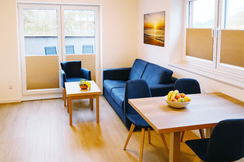 Living room interior of Fewos Haus am W&auml;ldchen with blue sofa, armchair, wooden tables, fruit bowls, and large glass doors and window