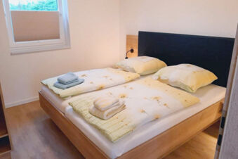 A double bed with floral bed linens, folded towels on each side, wooden floor, and a window at Fewos Haus am W&auml;ldchen