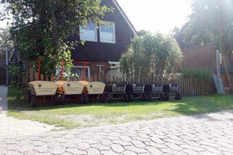 Several red-handled wagons with numbers are lined up on grass near a brick house at Fewos Haus am W&auml;ldchen