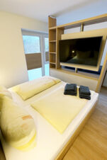 Bedroom with yellow bedding, black folded towels on the bed, mounted TV, wooden shelves, and window in Fewos Haus am W&auml;ldchen