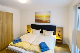 Bright bedroom at Fewos Haus am W&auml;ldchen with double bed, wooden wardrobe, bedside lamps, and a sunset beach picture on the wall