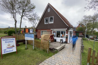 Two-story brown and brick building with wicker beach chair, signs, and a person walking on the path near Fewos Haus am W&auml;ldchen