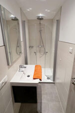 Small modern bathroom at Fewos Haus am W&auml;ldchen with glass shower, white sink, mirror, and orange towel on countertop